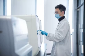 A person wearing a lab coat, face mask, safety glasses, and blue gloves operates a touchscreen panel on a large, white machine in a clean, bright laboratory setting focused on bioanalytical development.