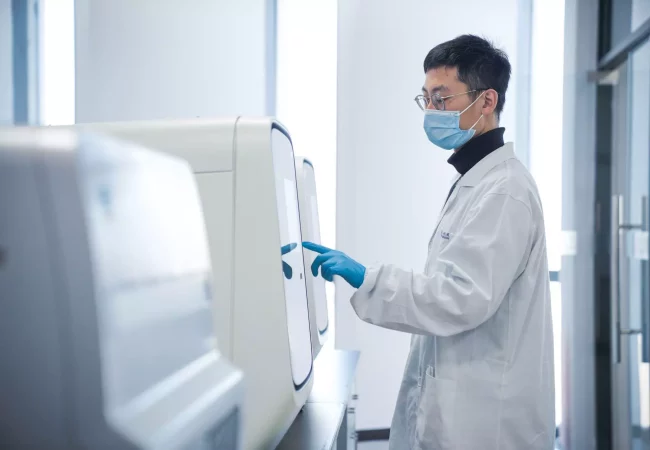 A person wearing a lab coat, face mask, safety glasses, and blue gloves operates a touchscreen panel on a large, white machine in a clean, bright laboratory setting focused on bioanalytical development.