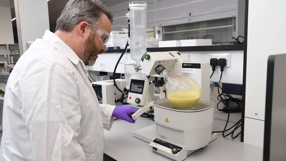A scientist wearing a lab coat and protective goggles is operating a rotary evaporator in a laboratory, meticulously observing a flask containing a yellow liquid. The lab, equipped for genetic toxicology research, has various tools and equipment on the counters and in the background.