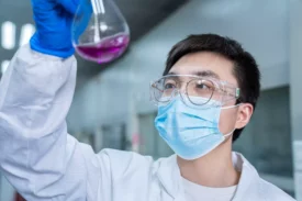 A scientist wearing a white lab coat, safety goggles, and a blue face mask holds up an Erlenmeyer flask containing a pink liquid. They are inside a laboratory, focused on the flask's contents while wearing blue gloves, conducting Preclinical ADME studies.