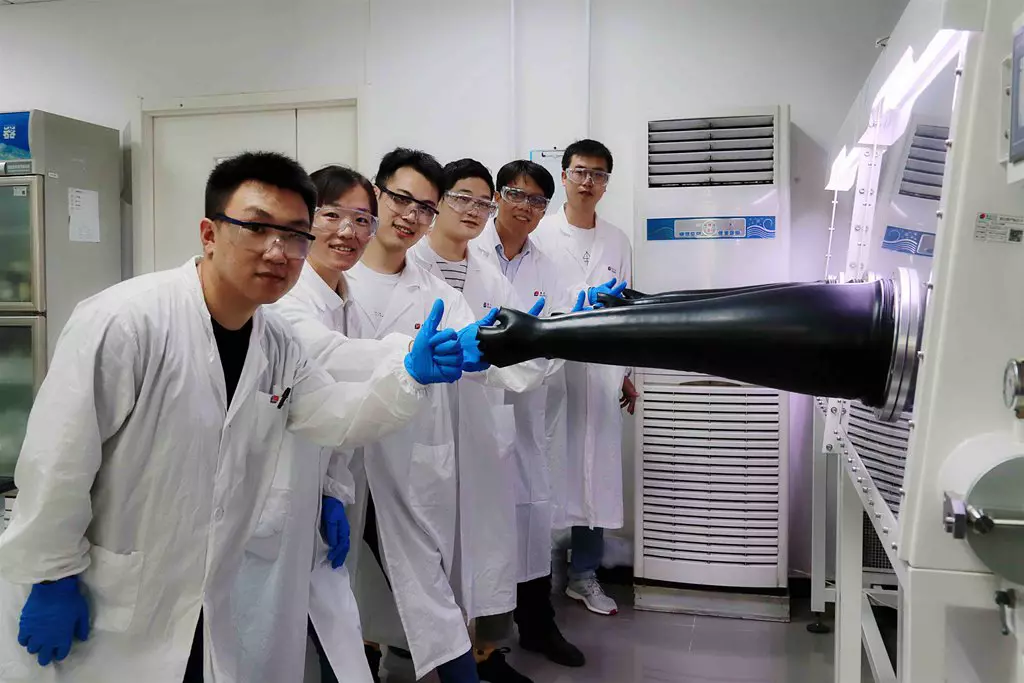 A group of six scientists in lab coats and safety glasses smile and gather around a glovebox, with one scientist demonstrating its use.