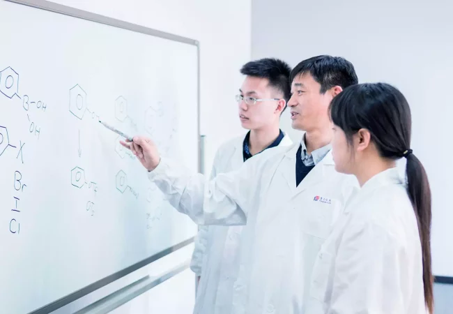 Three people in lab coats stand in front of a whiteboard covered with chemical formulas. A man in the middle, discussing bioanalytical development, points at one of the formulas with a marker, while the other two, a man and a woman, observe attentively.