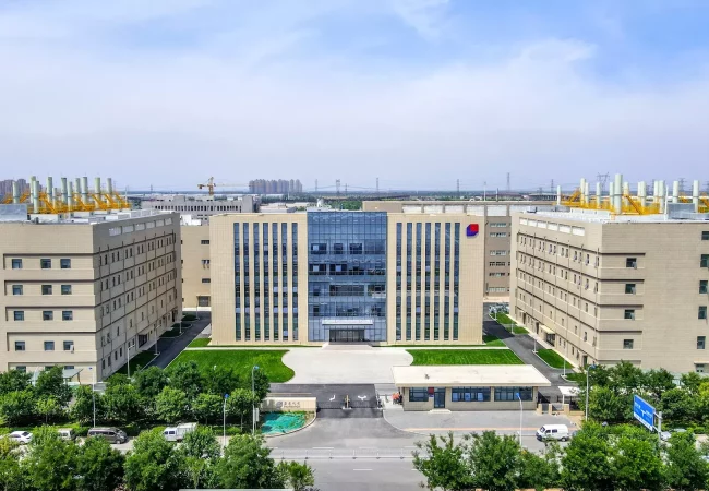 An aerial view of a modern office complex dedicated to bioanalytical development, featuring a central multi-story building flanked by two identical edifices. The facility is surrounded by trees and a well-maintained lawn, with clear skies and some distant industrial structures in the background.