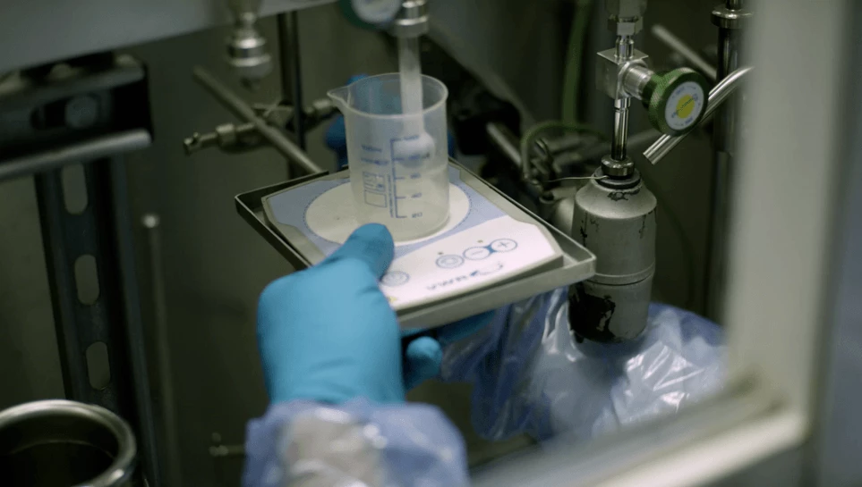 A scientist in blue gloves carefully measures a liquid in a graduated cylinder, performing tritium radiolabelling and radiosynthesis under controlled conditions.