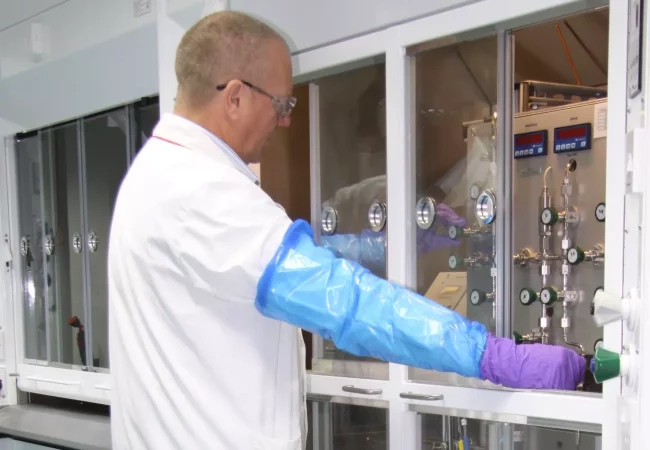 A scientist performs Tritium radiosynthesis in an enclosed lab setup, wearing long protective gloves and controlling precise instrumentation.