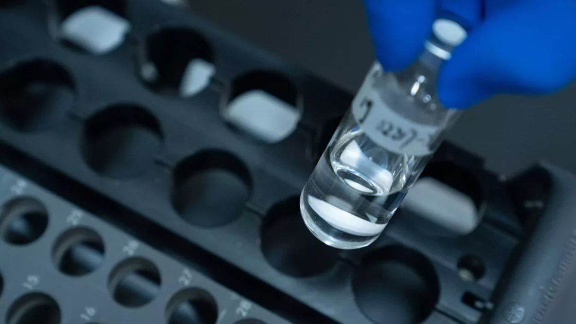 A gloved hand holds a small, clear vial filled with liquid, with numbers written on the label. The vial is being placed into or taken from a black, numbered holder with multiple circular compartments. This bioanalytical development setup is essential for precise measurements. The background is blurred.