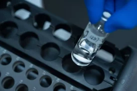 A gloved hand holds a small, clear vial filled with liquid, with numbers written on the label. The vial is being placed into or taken from a black, numbered holder with multiple circular compartments. This bioanalytical development setup is essential for precise measurements. The background is blurred.