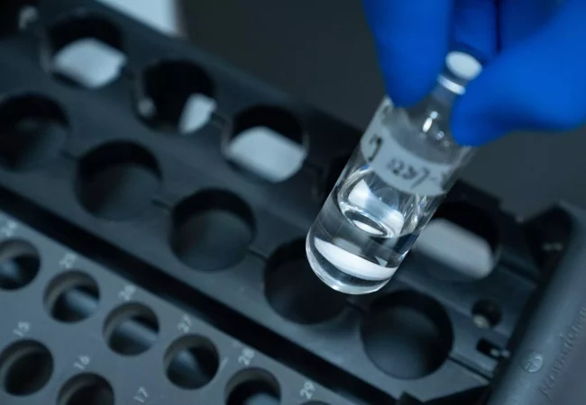 A gloved hand holds a small, clear vial filled with liquid, with numbers written on the label. The vial is being placed into or taken from a black, numbered holder with multiple circular compartments. This bioanalytical development setup is essential for precise measurements. The background is blurred.