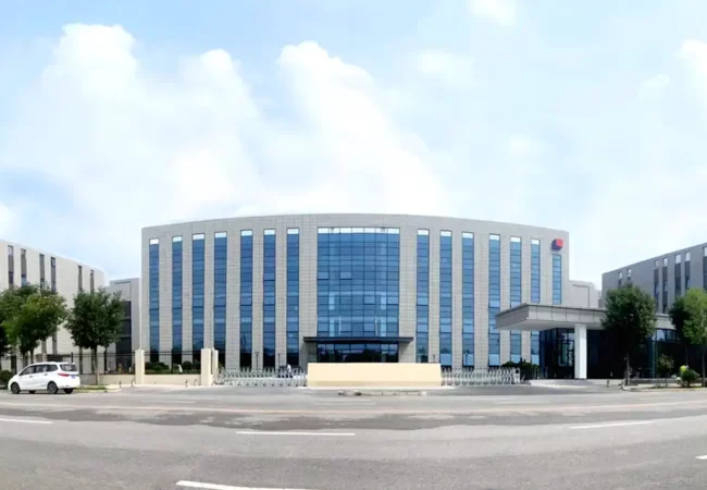 A modern, expansive office building with a sleek glass facade stands under a blue sky with scattered clouds. The building is surrounded by green trees, and a well-maintained garden area is in front. Several cars are parked on the street in front of the building that houses cutting-edge preclinical ADME studies labs.