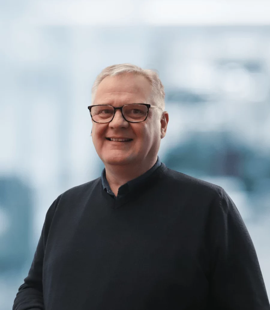 Tony Meaden, Associate Director, Quality Assurance & Core Team Leader, Pharmaron Gene Therapy. A man with short, light hair and glasses smiles warmly, wearing a dark sweater over a collared shirt. The background is softly blurred, suggesting an indoor office setting.