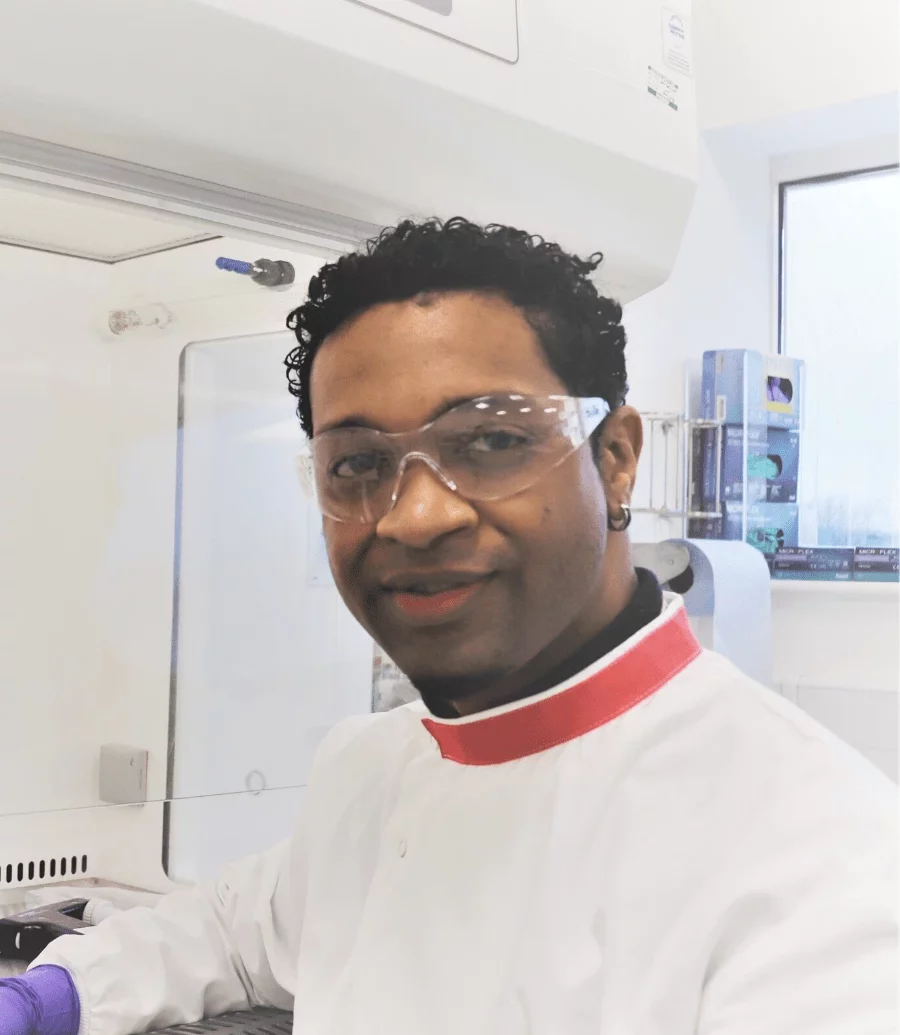 Shandel Pariag, Senior Scientist, Upstream Process Sciences, Pharmaron Gene Therapy - A male scientist with curly hair and safety glasses smiles in a laboratory. He wears a white lab coat with a red collar and purple gloves, working near scientific equipment in a well-lit environment.