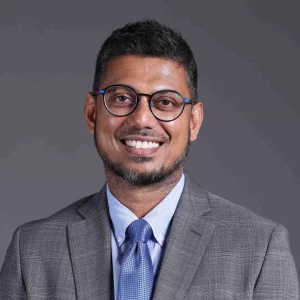 Sid Bhoopathy, Ph.D. - A man with short dark hair and a beard is smiling. He is wearing glasses, a light blue shirt, a blue tie, and a gray suit jacket. The background is plain and gray, akin to the sterile environment of preclinical ADME studies.