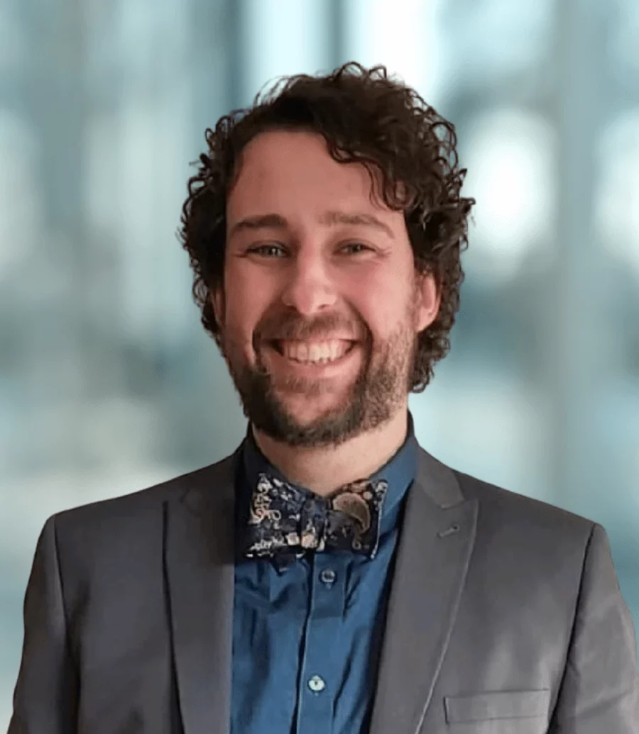 Siôn Edwards, Ph.D. - A man with curly hair and a beard smiles brightly, wearing a gray suit, blue shirt, and patterned bow tie. The background is softly blurred, suggesting a professional indoor setting.