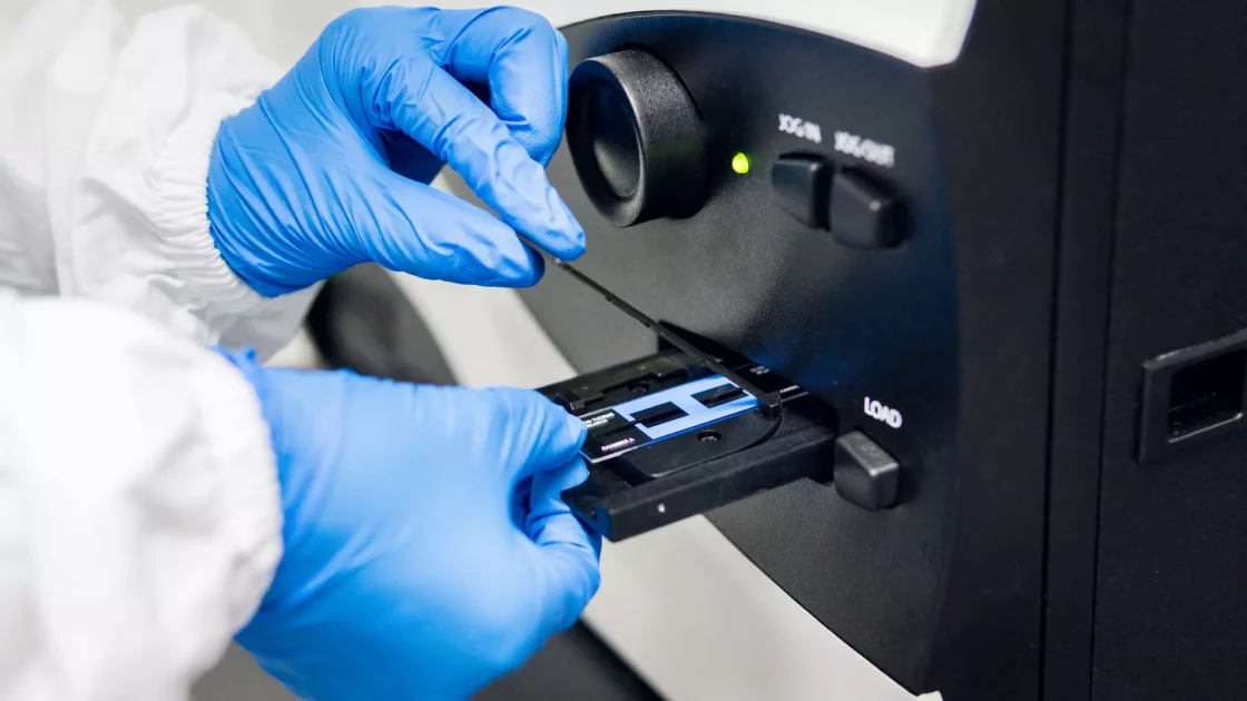 A scientist in protective gear inserts a sample slide into advanced lab equipment, likely for genetic toxicology analysis. The blue gloves and sterile environment highlight a focus on precision and contamination control.
