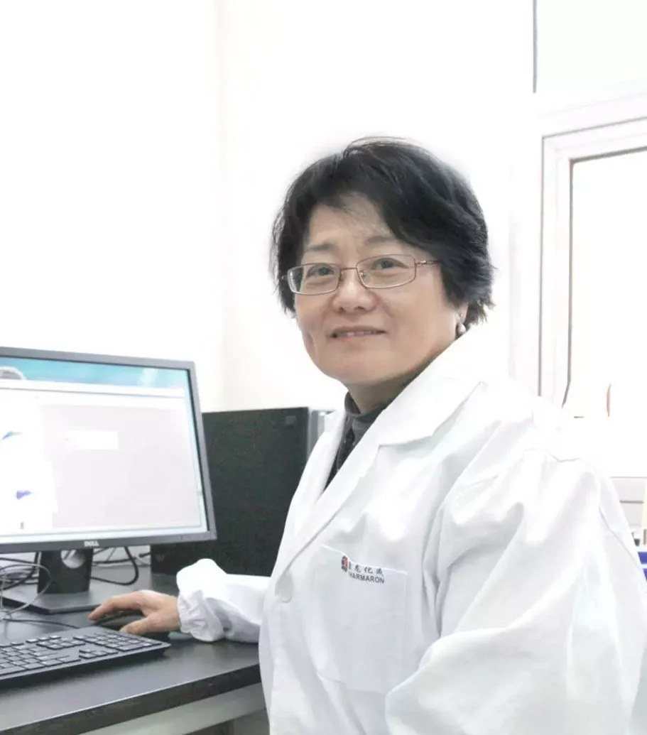 Jingqi Huang, PhD - A scientist with short black hair and glasses sits at a desk, working on a computer. Wearing a lab coat, she faces the camera, smiling in a bright, clean laboratory setting.
