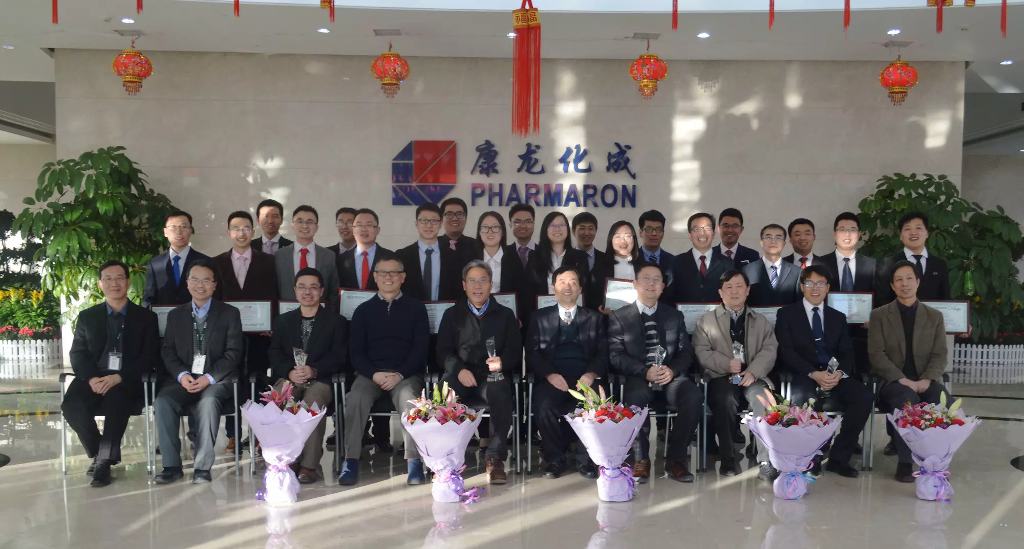 A group of professionals from the Pharmaron Leadership Class pose indoors with certificates, bouquets, and awards, under festive red lanterns.