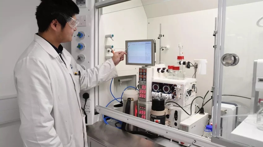 A scientist in a lab coat and protective glasses operates an automated reactor system, monitoring the process through a touchscreen interface in a laboratory setting.
