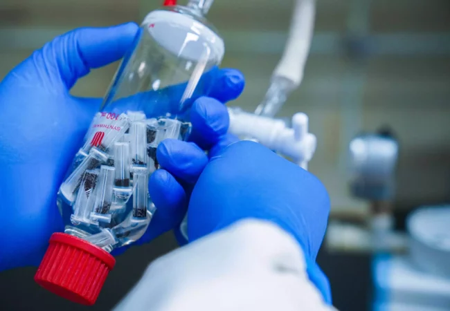 A scientist wearing blue gloves holds a bottle containing small vials used for carbohydrate analysis in a laboratory setting.