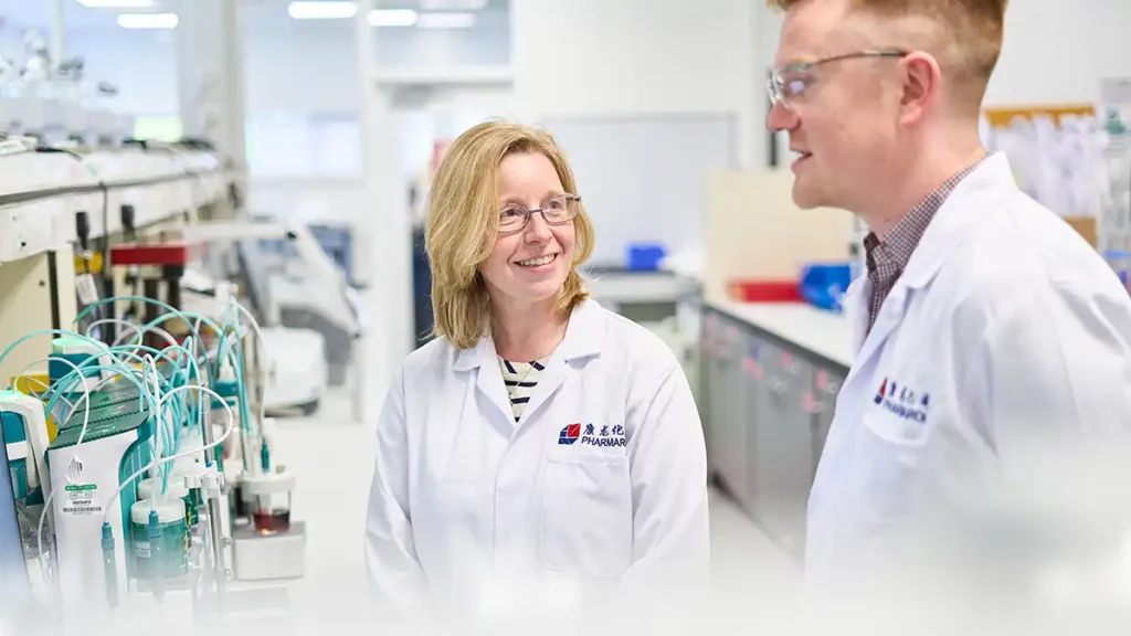 Two scientists in Pharmaron Cramlington lab discussing cGMP commercial manufacturing processes amid analytical lab equipment.