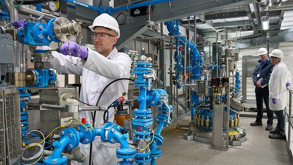 Scientist in PPE operates stainless steel equipment surrounded by blue pipelines in an advanced API manufacturing facility.