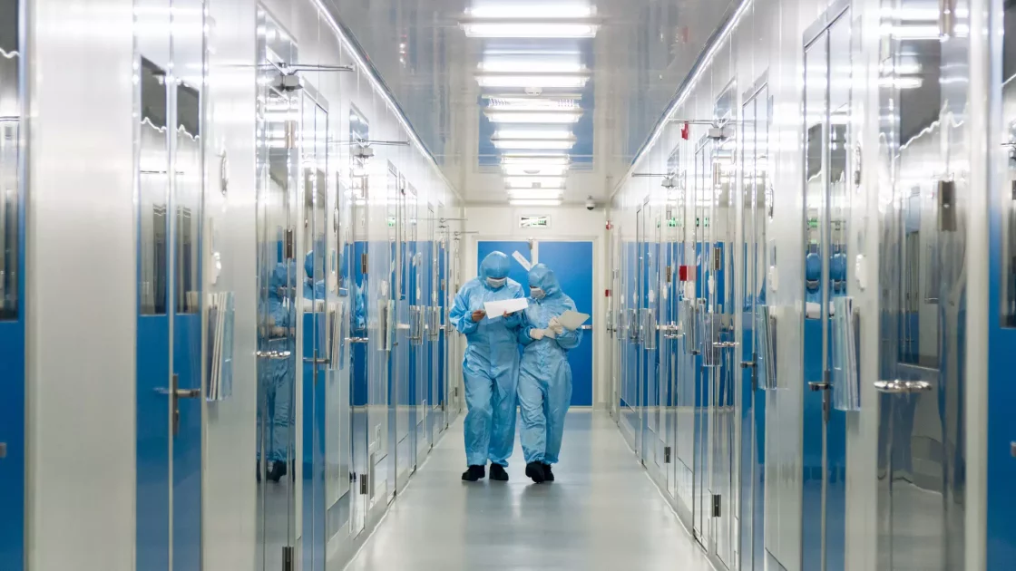 Two scientists in full protective suits review documents while walking through a cleanroom hallway in a sterile biopharma manufacturing facility.