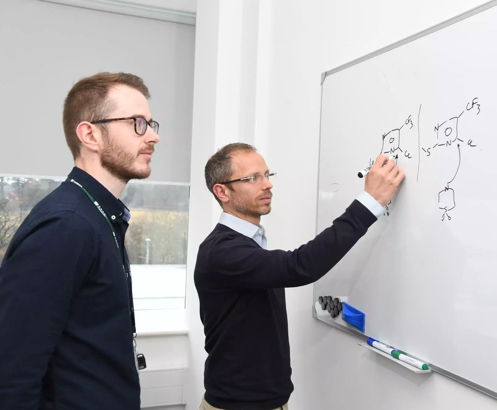 Two scientists discuss a chemical structure drawn on a whiteboard, likely related to medicinal chemistry research.