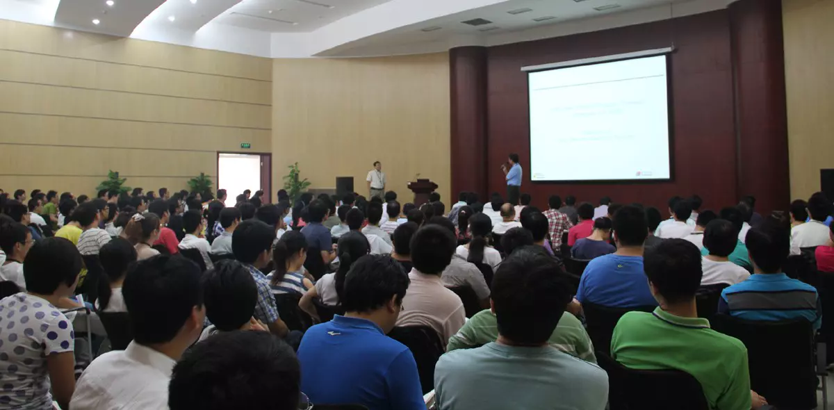 Large audience attending a Friday seminar with two speakers presenting in front of a projection screen in a spacious hall.