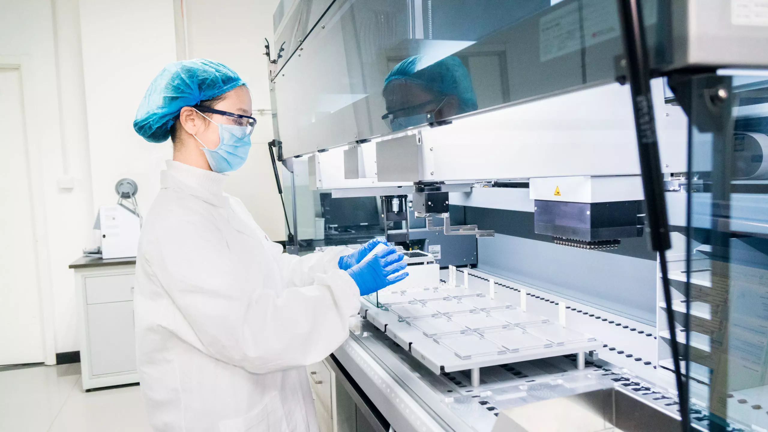 A scientist in protective gear operates an automated lab system for in vitro biology research, preparing biological samples for analysis.