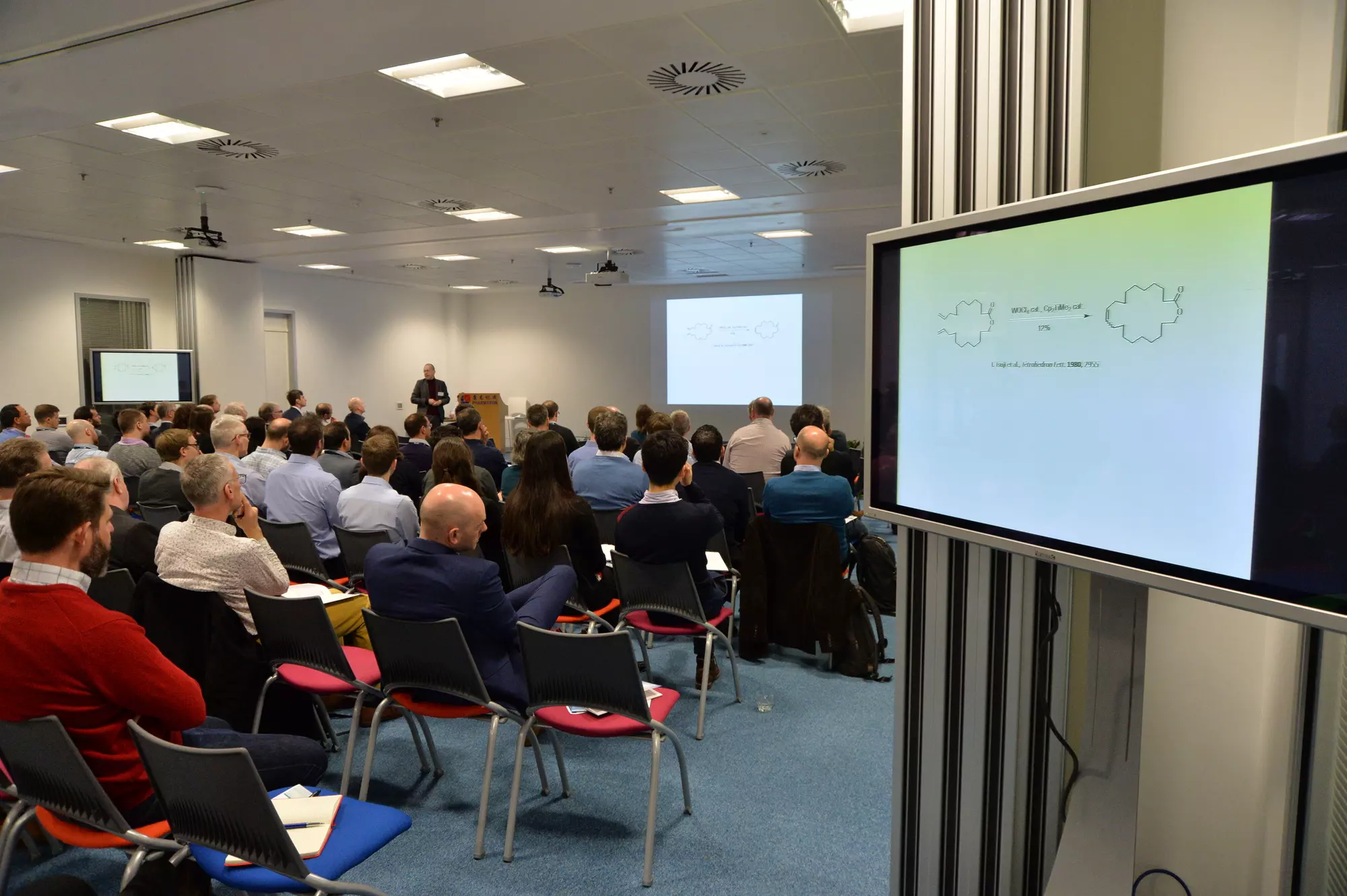 Pharmaron Science Seminar: A group of professionals attending a presentation in a conference room, with slides displayed on multiple screens showing chemical structures.