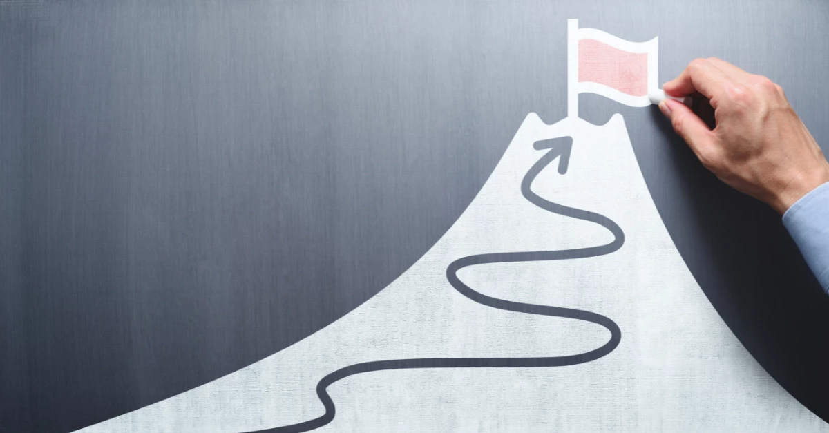 A hand places a red flag at the peak of a stylized mountain, with a winding path leading to the top. This visual represents achieving milestones, success, or reaching goals, symbolizing progress through challenges and strategic planning.