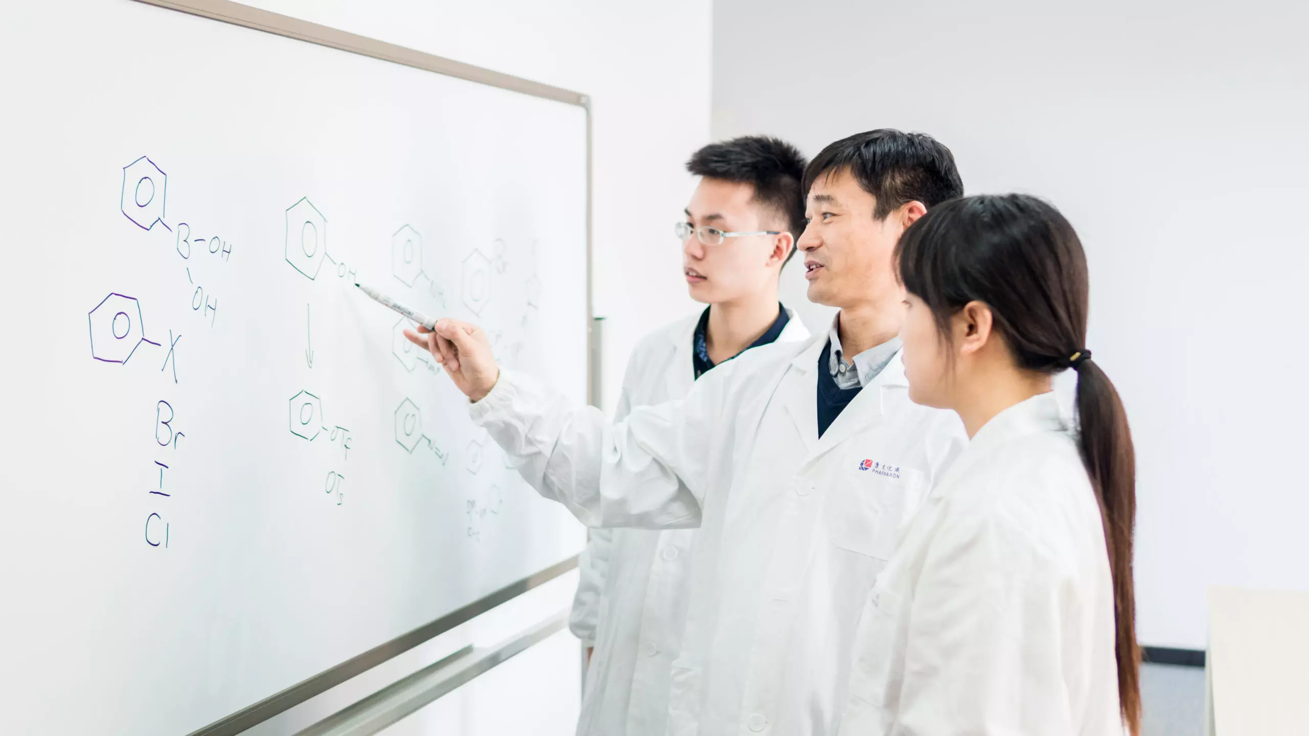 A scientist explains chemical structures on a whiteboard to two colleagues, discussing molecular diagrams and chemical reactions.