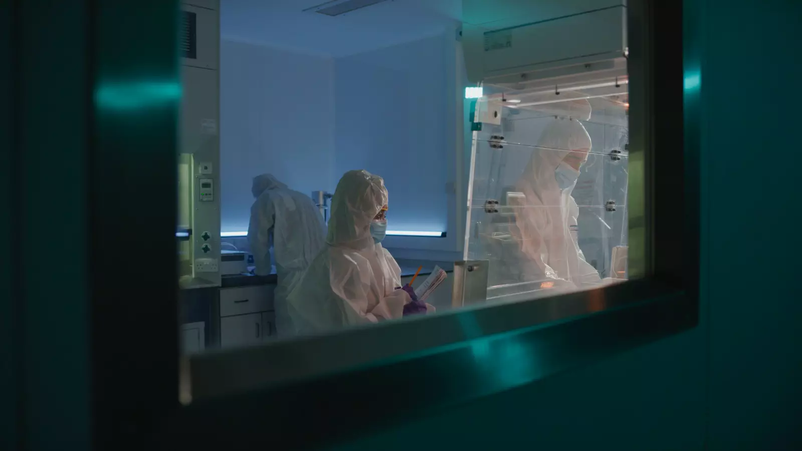 Scientists in full protective suits work inside a controlled lab environment at the Rushden facility, conducting radiolabeling experiments. One researcher documents data while another works under a fume hood.