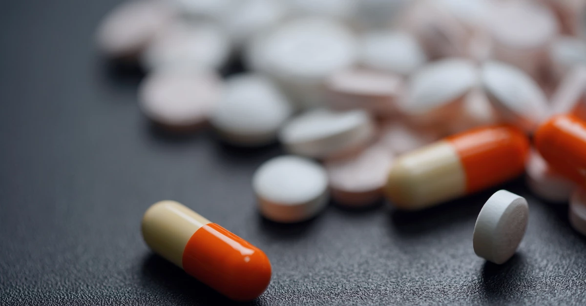 Close-up of various white round tablets and orange capsules scattered on a dark surface, with one capsule in the foreground.