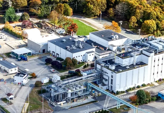 Aerial view of Pharmaron's Coventry site, a white multi-story facility with rooftop equipment, surrounded by trees with autumn foliage and an open driveway.