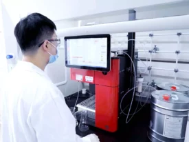 A lab technician wearing a white coat, safety goggles, and a face mask operates a machine with a large screen displaying data. Various tubes and containers, including a metal canister used in bioanalytical development, surround the equipment in a clean, sterile laboratory setting.