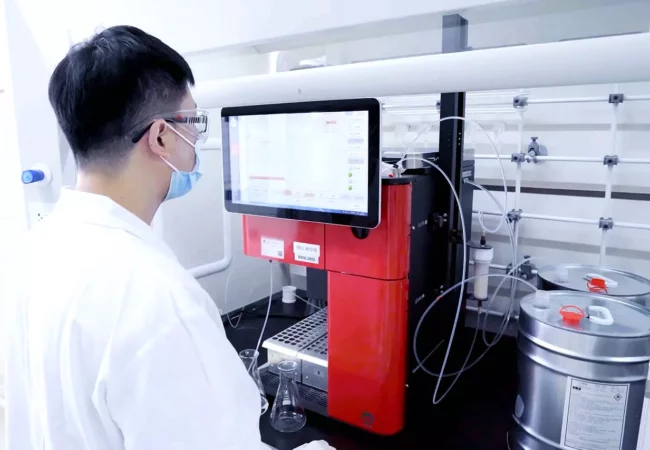 A lab technician wearing a white coat, safety goggles, and a face mask operates a machine with a large screen displaying data. Various tubes and containers, including a metal canister used in bioanalytical development, surround the equipment in a clean, sterile laboratory setting.