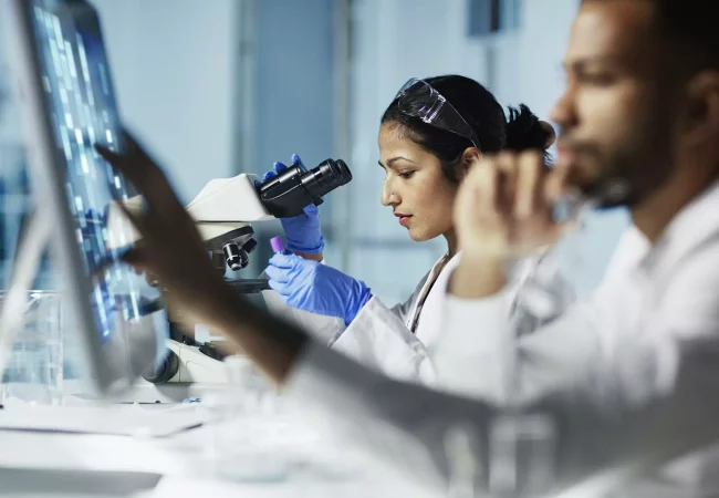 Scientists working in a lab, with one focusing on a microscope and another analyzing data on a screen, highlighting research and innovation.
