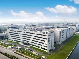 Aerial view of a modern industrial complex featuring multiple white-facade buildings, neatly landscaped green spaces, and several parked cars. The background shows a cloudy sky and additional industrial buildings where preclinical ADME studies are conducted.