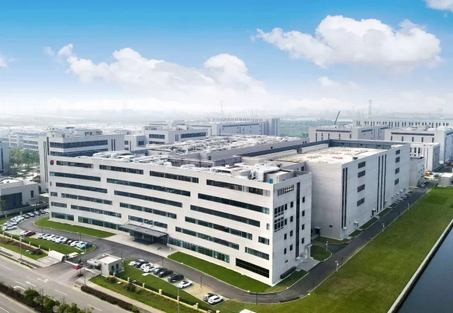 Aerial view of a modern industrial complex featuring multiple white-facade buildings, neatly landscaped green spaces, and several parked cars. The background shows a cloudy sky and additional industrial buildings where preclinical ADME studies are conducted.