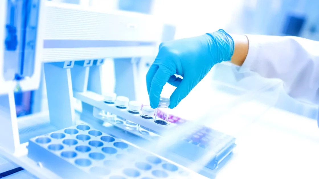 Gloved hand of a scientist placing a vial into an automated laboratory machine, surrounded by various sample trays in a brightly lit lab.