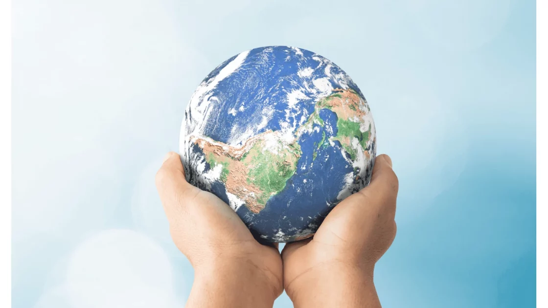 Two hands gently hold a small globe, symbolizing global social responsibility and environmental stewardship against a soft blue sky backdrop.