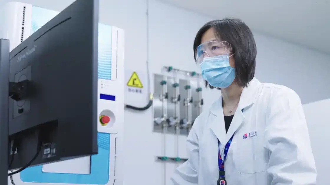Scientist in a Pharmaron lab coat, wearing safety goggles and a mask, analyzing data on a computer in a laboratory with advanced instrumentation.
