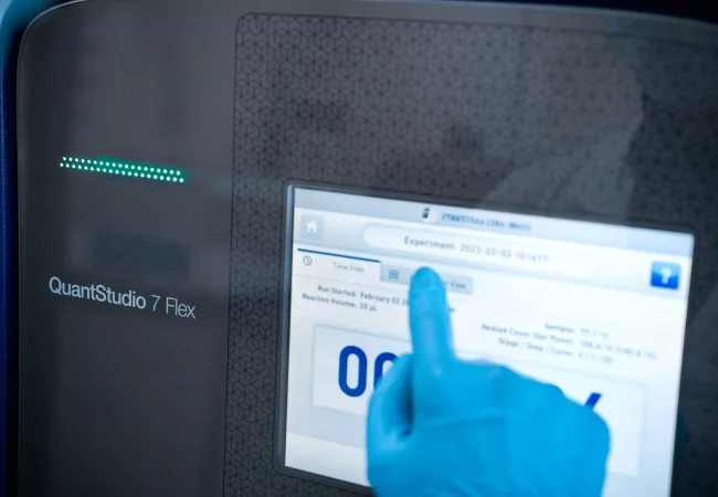 A gloved hand interacts with the touch screen of a QuantStudio 7 Flex instrument, displaying details of an ongoing biochemistry experiment.