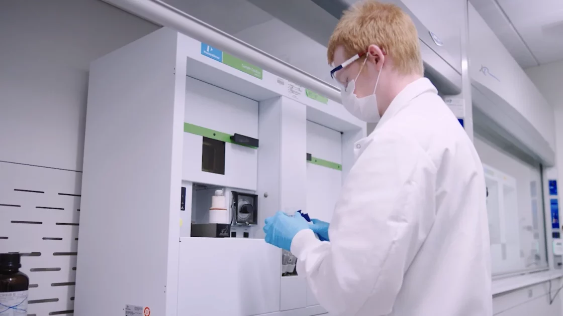 A scientist wearing protective gear handles a sample for Carbon-14 bioanalysis, using specialized lab equipment to measure isotope concentration.