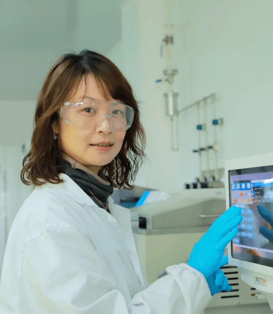 Jia Jia, Ph.D. A female scientist, wearing safety goggles, a lab coat, and blue gloves, operates a touchscreen in a laboratory. The background shows scientific equipment, emphasizing a controlled lab environment.