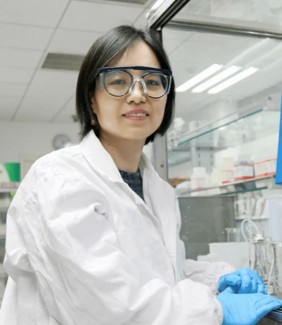 Mengxia Xue, Ph.D. A female scientist in safety goggles and a lab coat works in a laboratory. She smiles while wearing blue gloves, standing near lab equipment and glass containers on a clean workspace.