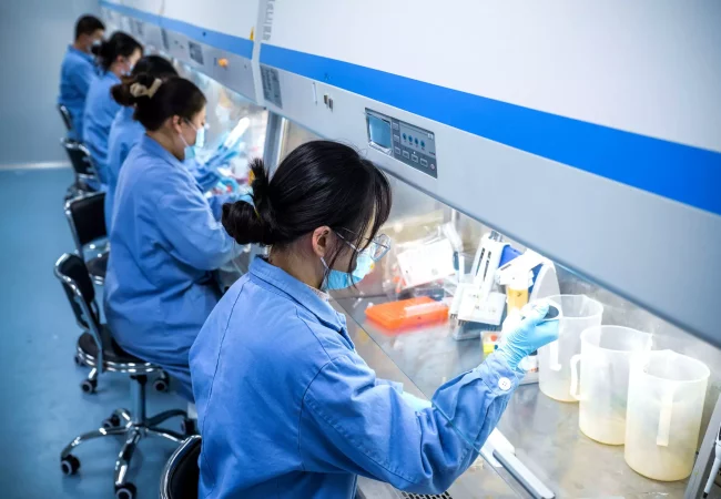 Scientists in blue lab coats and face masks are working in a row at laboratory benches, focusing on genetic toxicology. They use pipettes, containers, and other lab equipment under a sterile hood. The lab environment appears clean and organized.