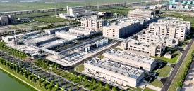 Aerial view of a large industrial complex specializing in genetic toxicology, with several beige buildings interconnected by pipes and walkways. The complex is surrounded by green fields, roads, and a rail line in the background. Trees and landscaped areas are visible within the compound.