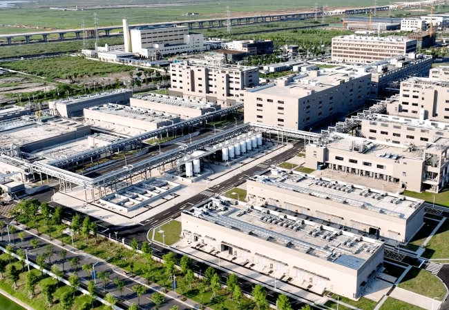 Aerial view of a large industrial complex specializing in genetic toxicology, with several beige buildings interconnected by pipes and walkways. The complex is surrounded by green fields, roads, and a rail line in the background. Trees and landscaped areas are visible within the compound.