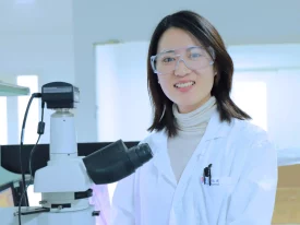 A smiling scientist in protective eyewear stands beside a microscope in a lab, representing CGT ophthalmology research and development.